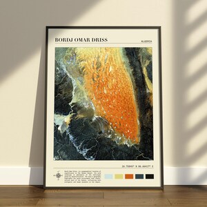 Op de afbeelding: Een ingelijste kunstdruk met een satellietweergave van Bordj Omar Driss, Algerije. De afbeelding toont oranje woestijnduinen in contrast met donker terrein. De tekst bevat de plaatsnaam, coördinaten en een kleurenpalet met vijf stalen.