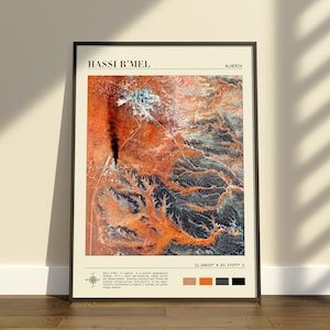 Op de afbeelding: Een verticale wandkunstprint met een satellietweergave van Hassi R'Mel, Algerije. De kaart toont oranje woestijnterrein met donkere geologische formaties, zwarte tekstlabels, coördinaten en een klein kleurenpalet van oranje, grijs en zwart.
