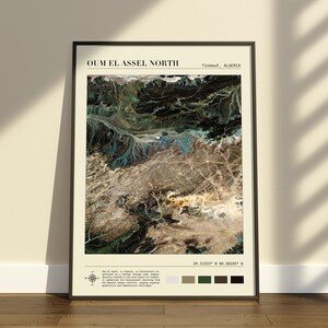 Op de afbeelding: Een ingelijste wandkunstprint met een satellietweergave van Oum El Assel North, Tindouf, Algerije. De afbeelding toont woestijnterrein met donkere, getextureerde geologische formaties en lichte zandgebieden. Tekst bevat coördinaten en notities.