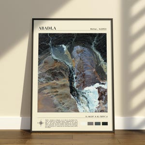 Op de afbeelding: Een ingelijste wandkunstprint met een satellietweergave van Abadla, Béchar, Algerije. De kaart toont een contrastrijk luchtfoto-landschap met donkere, kronkelende rivierbeddingen, lichtgekleurd woestijnterrein en tekst met coördinaten.