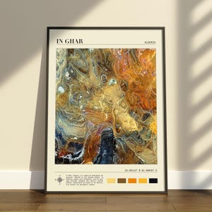 Op de afbeelding: Een ingelijste kunstdruk getiteld IN GHAR ALGERIA met een satellietachtig luchtfoto-aanzicht van woestijnterrein met wervelende patronen in bruin, goud, blauw en zwart. De print bevat coördinaten, een windroos en kleurstalen.