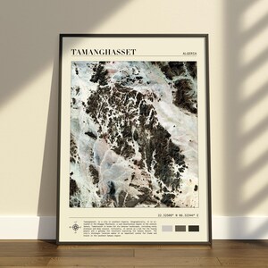 Könnte beinhalten: Ein gerahmter Kunstdruck mit einer Satellitenansicht von Tamanghasset, Algerien. Das Bild zeigt kontrastreiches felsiges Gelände in Schwarz-, Weiß- und Beigetönen. Der Text enthält den Stadtnamen, Koordinaten und einen beschreibenden Text.