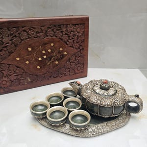 May include: A dark green stone tea set featuring a teapot with a silver-toned metal overlay, six matching small cups, and an oval silver-toned tray. A rectangular wooden box with intricate floral carvings and gold-toned inlay sits behind.