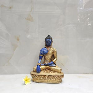 May include: A seated Buddha statue featuring a deep blue finish on the head, torso, and arms, contrasted with a gold-toned robe and base. The figure holds a small clear sphere in one hand and is accompanied by a single white and yellow flower.