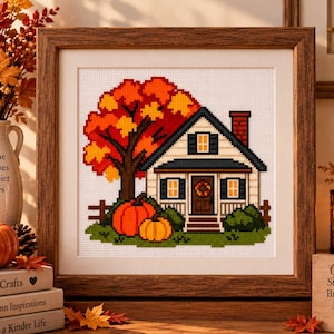 May include: A framed cross-stitch artwork depicting a house with a red brick chimney, a large autumn tree with orange and yellow leaves, and two orange pumpkins on green grass. The house features dark shutters, a front door with a wreath, and a porch.