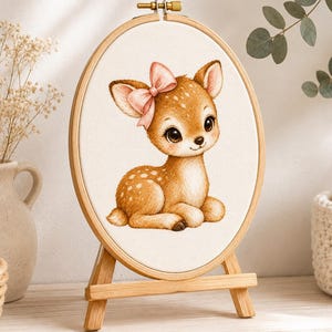 Peut inclure: Un tambour à broder ovale en bois contenant un panneau en tissu avec une illustration imprimée d'un faon brun à taches blanches portant un nœud rose sur la tête. Le tambour est monté sur un petit chevalet en bois, présentant l'œuvre debout.