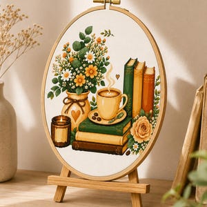 May include: An oval embroidery hoop on a wooden stand featuring a stitched illustration of a yellow coffee mug on a stack of books, a vase with yellow and white flowers, a lit candle, and a small heart, all rendered in warm, earthy tones.