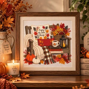 May include: A framed cross-stitch artwork depicting a wooden bench with a pillow reading Enjoy the little things, a plaid blanket, a mug, a lantern, and pumpkins. The scene features autumn leaves in shades of orange, red, and yellow on a white fabric.