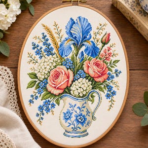 May include: An oval embroidery hoop containing a cross-stitch pattern of a floral bouquet in a blue and white pitcher. The arrangement features a large blue iris, two pink roses, white hydrangea clusters, and small blue flowers on a cream fabric.