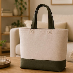 May include: A structured tote bag featuring a two-tone design with a cream-colored upper section and a dark olive green base. The bag includes two matching dark olive green fabric handles securely attached with reinforced stitching at the top.