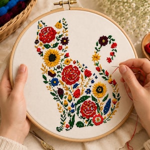 May include: A round wooden embroidery hoop containing white fabric with a cross-stitch design of a cat silhouette filled with colorful floral patterns including red roses, yellow sunflowers, and green leaves, with a needle and red thread.