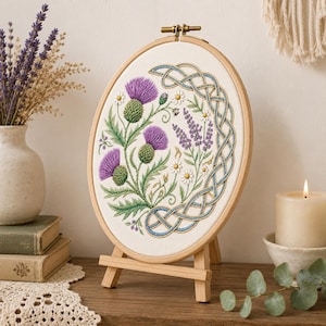 Puede incluir: Un bastidor de bordado ovalado que contiene un panel de tela color crema con un diseño detallado de flores de cardo moradas, pequeñas margaritas blancas y ramitas de lavanda, enmarcado por un intrincado patrón de nudo celta azul y dorado.