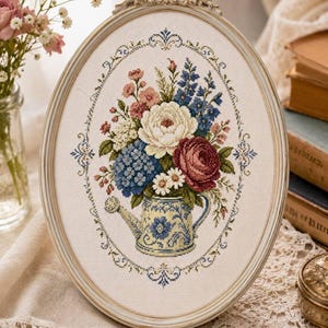 May include: An oval-framed cross-stitch embroidery featuring a bouquet of white, pink, blue, and deep red flowers arranged in a blue and white patterned watering can. The floral design is surrounded by a delicate blue and gold decorative border.