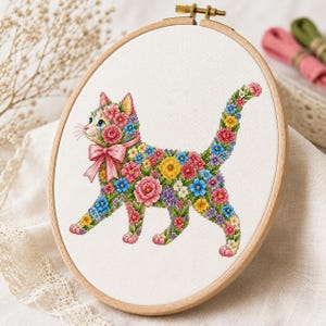 Op de afbeelding: Een borduurring met witte stof en een kruissteekontwerp van een kat die volledig uit kleurrijke bloemen bestaat. De kat heeft een roze strik om de nek en is gevuld met diverse kleine blauwe, gele en roze bloemmotieven.