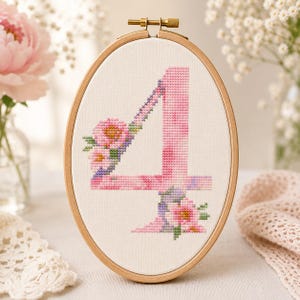 May include: An oval wooden embroidery hoop containing white fabric with a cross-stitched number four. The number is rendered in shades of pink and purple, decorated with small cross-stitched floral clusters in pink, green, and yellow hues.