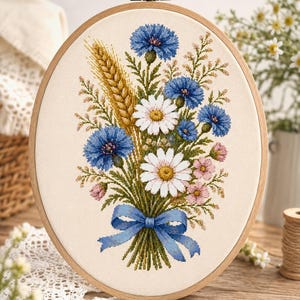Pode incluir: Um bastidor de bordado oval de madeira contendo um padrão de ponto cruz de um buquê de flores. O design apresenta centáureas azuis, margaridas brancas, flores rosas e espigas de trigo douradas, atadas na base com um laço de fita azul.