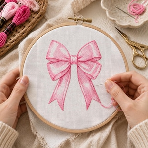 May include: A circular wooden embroidery hoop containing white Aida fabric with a cross-stitched design of a large, vibrant pink bow. The bow features detailed shading in various shades of pink, creating a dimensional appearance on the white cloth.