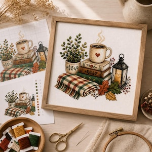 May include: A framed cross-stitch embroidery featuring a stack of three books topped with a steaming mug with a heart, a potted plant, a plaid blanket, a lit lantern, and autumn leaves. The design is displayed on white fabric in a light wood frame.