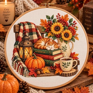 Puede incluir: Un bastidor de bordado circular con un diseño de punto de cruz de un gato atigrado naranja durmiendo sobre una pila de libros. La escena incluye una linterna encendida, una calabaza, una taza de café humeante y un ramo de girasoles y hojas de otoño.