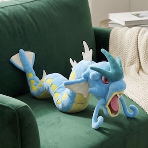 May include: A blue plush toy shaped like a serpentine creature with a long body, yellow circular markings along its sides, white fins, and a large head featuring a wide-open mouth with a pink tongue, sharp teeth, and prominent teal horns.