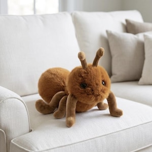 Peut inclure: Une peluche en forme de fourmi faite d'un tissu marron doux et duveteux. L'insecte en peluche présente une tête arrondie avec deux antennes dressées, de grands yeux sombres et six pattes articulées s'étendant depuis son corps.
