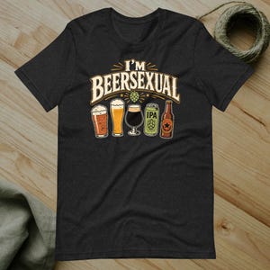 May include: A charcoal gray short-sleeve t-shirt featuring the text I'M BEERSEXUAL in a cream-colored font. Below the text are illustrations of three different beer glasses, a green IPA beer can, and a brown beer bottle with a star label.