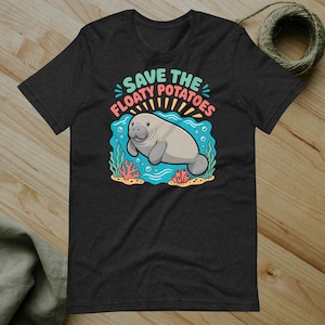 May include: A black short-sleeve t-shirt featuring a graphic of a grey manatee swimming in blue water with coral and bubbles. The text above the manatee reads "SAVE THE FLOATY POTATOES" in light blue and pink lettering, centered on the chest.
