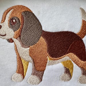 Pode incluir: Um design bordado de um filhote de beagle em um fundo de tecido branco. O filhote apresenta pelagem marrom, bege e cinza escuro com peito e focinho brancos. O cão está de perfil, voltado para a esquerda, com a cauda apontada para cima.