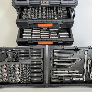 May include: A black Ridgid tool storage system with two open drawers containing various silver and black metal sockets, extensions, and wrenches. The lower section features two side-by-side trays holding Husky brand tools, including hex keys, impact sockets, and ratchets organized in molded plastic compartments.