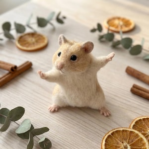Könnte beinhalten: Eine nadelgefilzte Hamsterfigur aus cremefarbenen und hellbraunen Wollfasern mit kleinen schwarzen Perlenaugen, einer rosa Nase und ausgestreckten Vorderpfoten. Der Hamster steht aufrecht auf seinen Hinterbeinen in einer dynamischen Pose.
