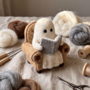 Puede incluir: Una figura de fantasma blanco de lana fieltrada con ojos de cuentas negras, sentada en un sillón color canela, sosteniendo un libro gris abierto. El fantasma tiene una textura suave y difusa, posicionado como si estuviera leyendo en la silla.