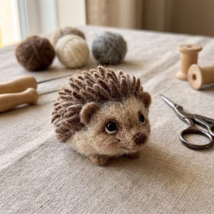 Op de afbeelding: Een naaldgevilte egelbeeldje gemaakt van bruine en beige wolvezels. De egel heeft een getextureerde, stekelige rug, een rond gezicht met donkere kraaloogjes en een kleine snuit. Het is geplaatst tussen diverse knutselbenodigdheden.