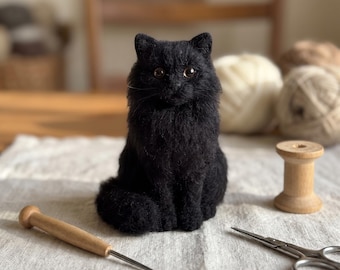 Op de afbeelding: Een naaldgevilte zwarte katfiguur met realistische bruine ogen en een getextureerde vacht. De kat zit rechtop en kijkt naar voren. Het beeldje is gemaakt van fijne wolvezels en geeft het uiterlijk van een langharige kat weer.