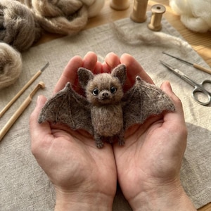 May include: A small, brown, needle-felted bat figurine with large ears, dark eyes, and outstretched wings. The bat is crafted from textured wool fibers, featuring a rounded body, pointed ears, and detailed wing membranes.