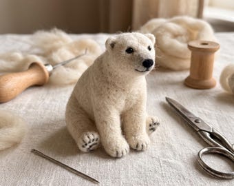 Può includere: Una statuetta di orso polare in lana infeltrita color bianco sporco, con piccoli occhi neri, un naso nero e cuscinetti delle zampe ricamati. L'orso è circondato da articoli per il fai da te, tra cui un attrezzo in legno, un ago e forbici.