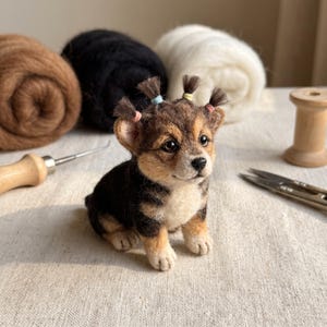 May include: A needle-felted puppy figurine with brown, black, and cream fur. The puppy has three small tufts of hair on its head secured with tiny pink, blue, and yellow elastic bands. It features dark eyes, a black nose, and a seated posture.