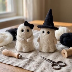 Puede incluir: Dos figuras de fantasmas de lana fieltrada blanca con ojos de cuentas negras. Un fantasma lleva un pequeño lazo negro y el otro un sombrero puntiagudo negro. Un punzón con mango de madera y unas tijeras de metal descansan sobre una tela clara.