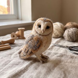 May include: A needle-felted barn owl figurine with a heart-shaped face, black eyes, and textured plumage in shades of cream, tan, and grey. The owl stands on small, detailed feet, crafted from wool fibers with a soft, fuzzy appearance.