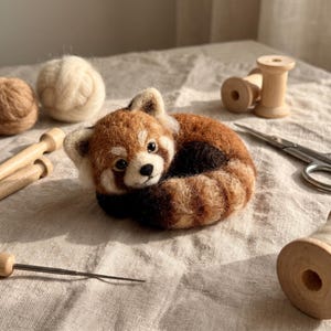 May include: A needle-felted red panda figurine with a curled tail, featuring a mix of rust-orange, dark brown, and cream-coloured wool fibres. The small animal has black bead eyes, a black nose, and pointed ears with white inner fur details.