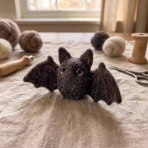 Könnte beinhalten: Eine dunkelbraune, nadelgefilzte Fledermausfigur mit rundem Körper, spitzen Ohren und ausgebreiteten Flügeln. Die Fledermaus hat zwei große, glänzende, helle Augen, die in ihre flauschige, texturierte Wolloberfläche eingearbeitet sind und detailliert wirken.