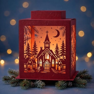 May include: A deep red paper lantern featuring intricate laser-cut silhouettes of a church, small houses, and pine trees. The design includes a crescent moon and stars, with a warm internal light illuminating the cutouts from within.