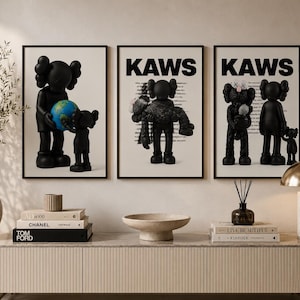 Op de afbeelding: Een set van drie ingelijste wandposters met zwarte KAWS-figuren. De eerste poster toont twee figuren die een wereldbol vasthouden, terwijl de tweede en derde poster KAWS-figuren met tekst op een witte achtergrond weergeven.