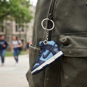 May include: A miniature high-top sneaker keychain featuring a two-tone navy blue and light blue color scheme with a white midsole. The side of the shoe displays a navy blue swoosh logo and a circular graphic with the text Hoops College.