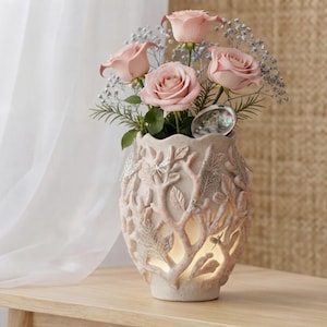 May include: A light pink ceramic vase featuring a raised, textured branch and leaf design with integrated lighting. The vase holds a bouquet of three soft pink roses, sprigs of baby's breath, and a single iridescent abalone shell.