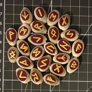Puede incluir: Una colección de veinticuatro piedras blancas de forma ovalada, cada una con una incrustación circular lisa de color rojo oscuro pintada con un símbolo rúnico dorado único. Las piedras están dispuestas en un grupo sobre una superficie negra cuadriculada.