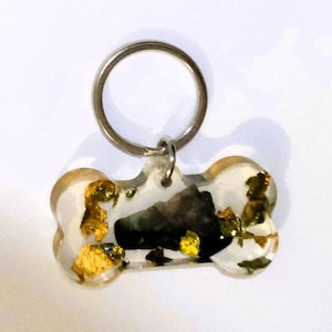 Puede incluir: Una placa para mascota con forma de hueso hecha de resina transparente, con copos de pan de oro incrustados y un fragmento central oscuro de textura similar a la piedra. La placa está unida a una anilla metálica plateada para el collar.