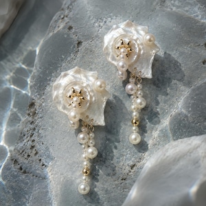 May include: A pair of earrings featuring iridescent white seashell bases, each adorned with a central gold-toned star-shaped accent and multiple dangling white pearls. The design incorporates small gold-toned beads interspersed among the pearls.