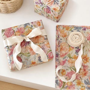 Puede incluir: Una caja de regalo cuadrada envuelta en papel floral con un lazo de cinta color crema, junto a una pila de hojas de papel de regalo a juego y un rollo de cinta color crema, todo con un estampado botánico estilo acuarela.