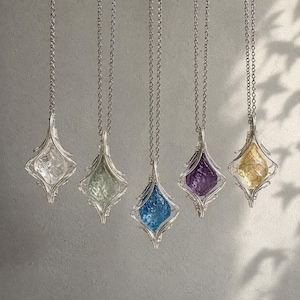 May include: Five necklaces featuring raw, faceted gemstones encased in silver-toned wire cages. The pendants include clear, light green, blue, purple, and yellow stones, each suspended from a matching silver-toned chain in a vertical arrangement.