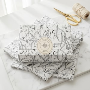 May include: A rectangular gift box wrapped in white paper with a black line-art floral pattern. A circular beige sticker on the centre features the text "HAPPY MOTHER'S DAY" and "Best wishes". Gold-toned metal scissors and a spool of twine are nearby.